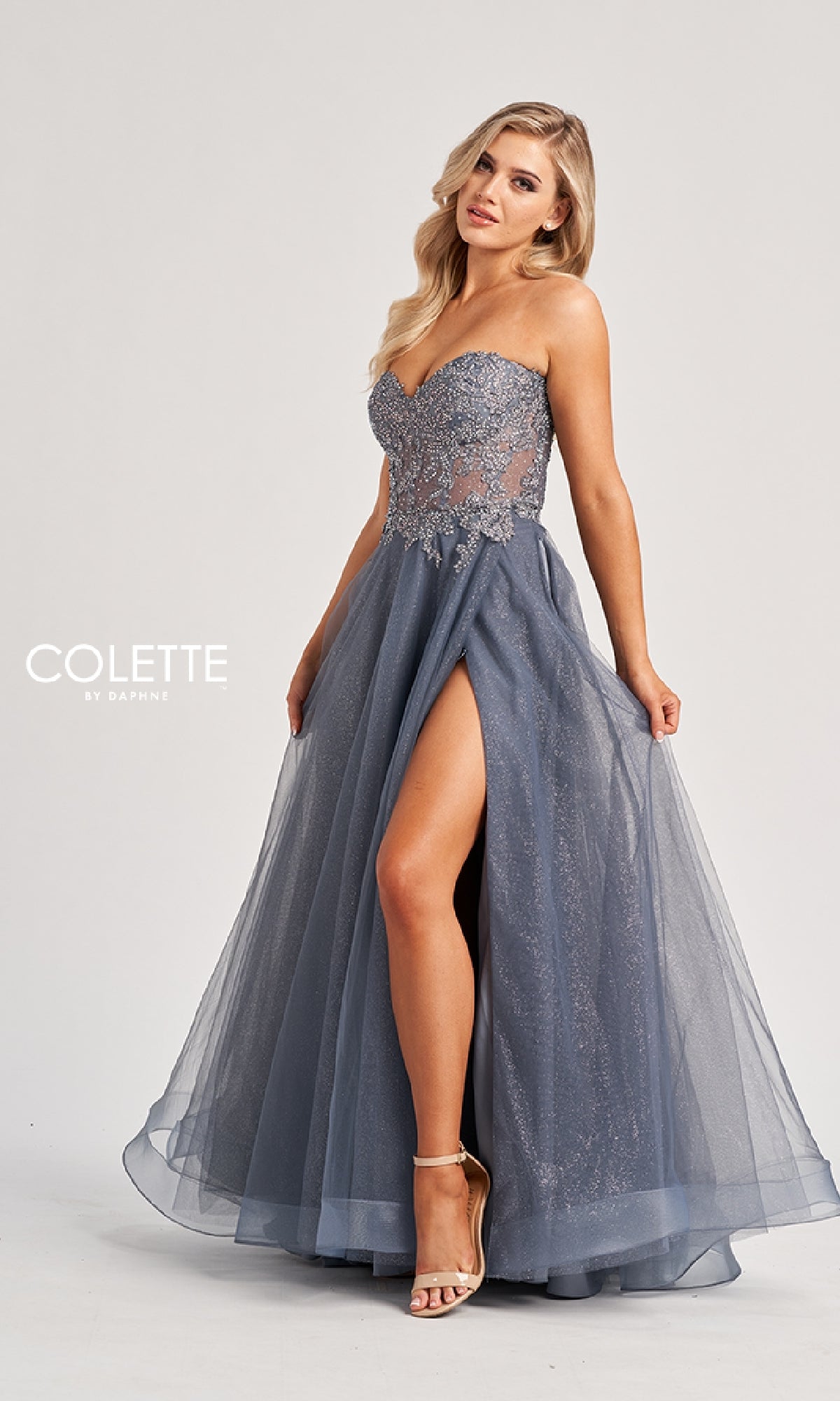 A woman wears the Long Prom Dress: Colette CL8110, featuring a strapless embroidered corset bodice and a blue-gray, floor-length A-line skirt with a high slit, standing against a plain background.